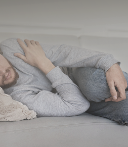Man lying curled up on a sofa, representing fatigue, discomfort, and the need for rest due to health issues