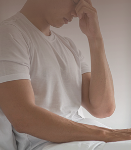 Man sitting on the edge of the bed with his head in his hand, representing despair, stress, or the psychological burden of a health condition