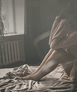 Woman sitting curled up on the bed with head on knees, representing isolation and emotional distress