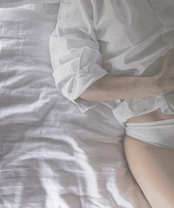 Overhead close-up of a woman lying on a bed in white underwear with her hand on her abdomen, representing intimate health, hygiene, and body awareness